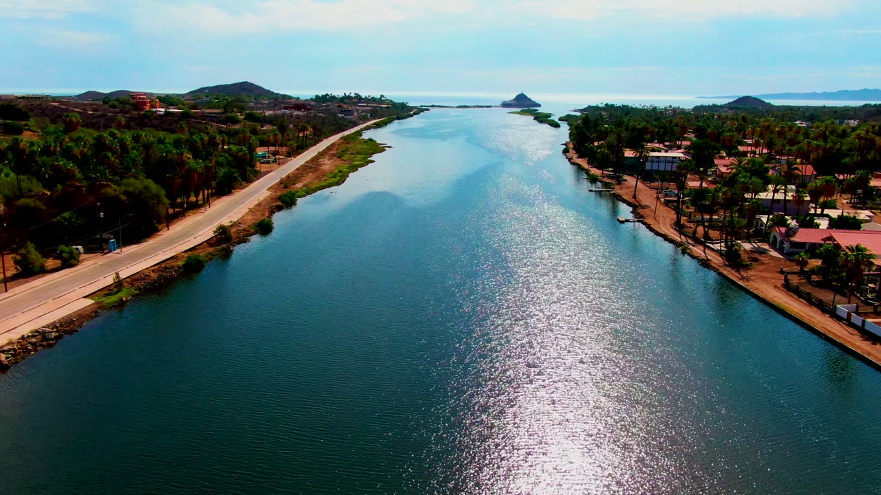Opening aerial frame over Mulege, Baja California Sur, with a blue-green canal centered between a paved road, palms, houses, distant hills, and no visible people.