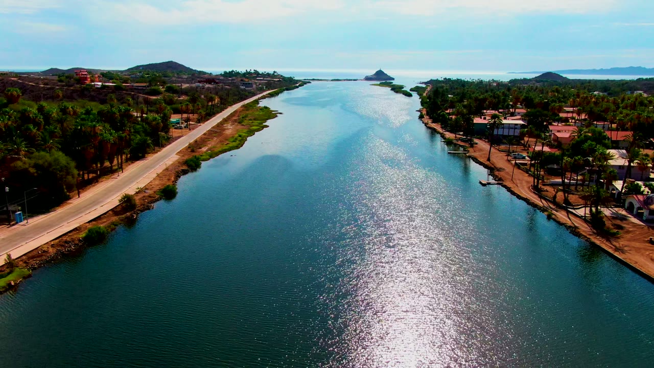 Mid-clip aerial view in Mulege with the water glare widening in the canal, palms and homes along the right side, road on the left side, and no visible people.