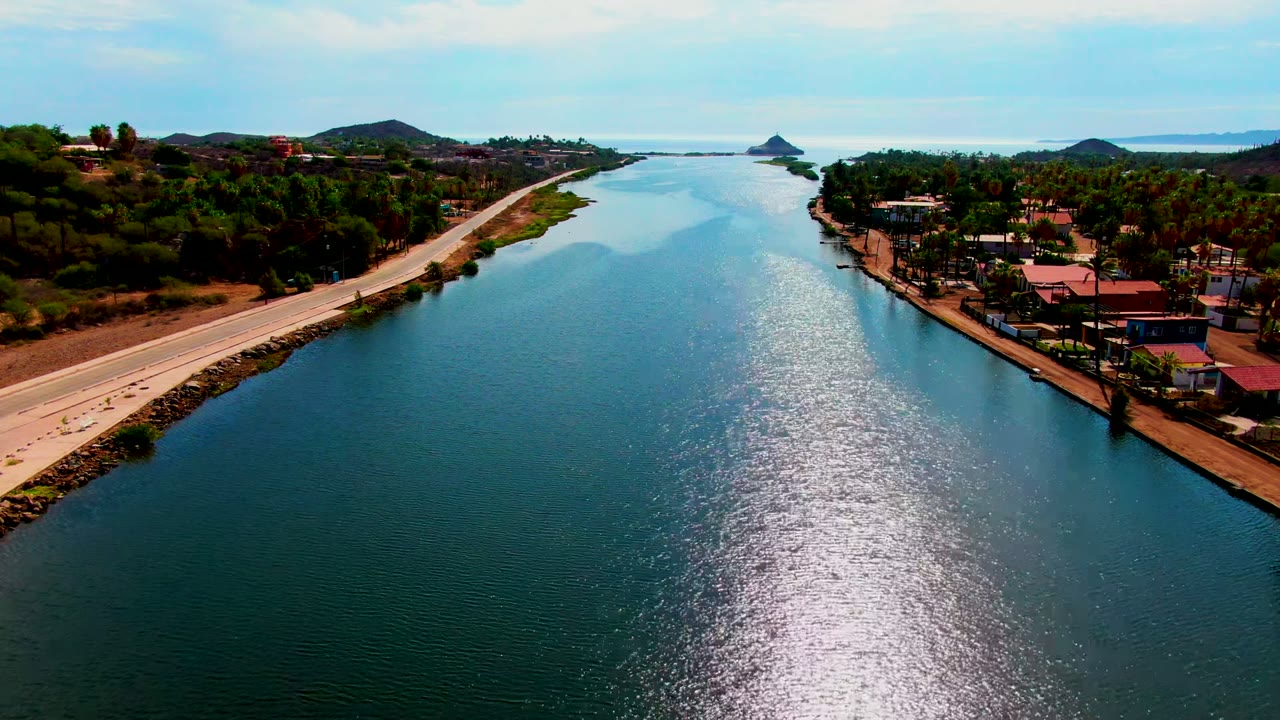 Opening aerial view in Mulege, Baja California Sur, Mexico, with a road along the left bank, a broad reflective waterway in the center, red-roofed homes and palms on the right, low hills beyond, and no visible people.
