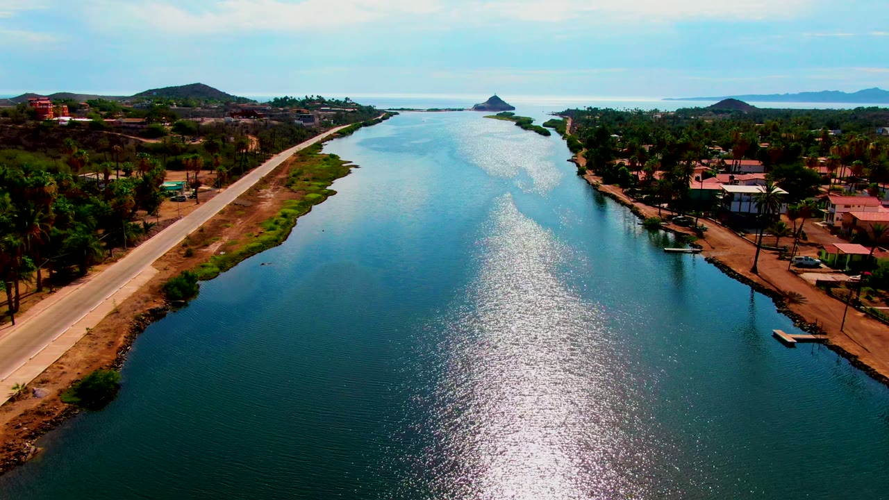 Opening aerial view in Mulege, Baja California Sur, showing a canal between a paved road and shoreline buildings with palms, distant hills, bright reflected water, and no visible people.