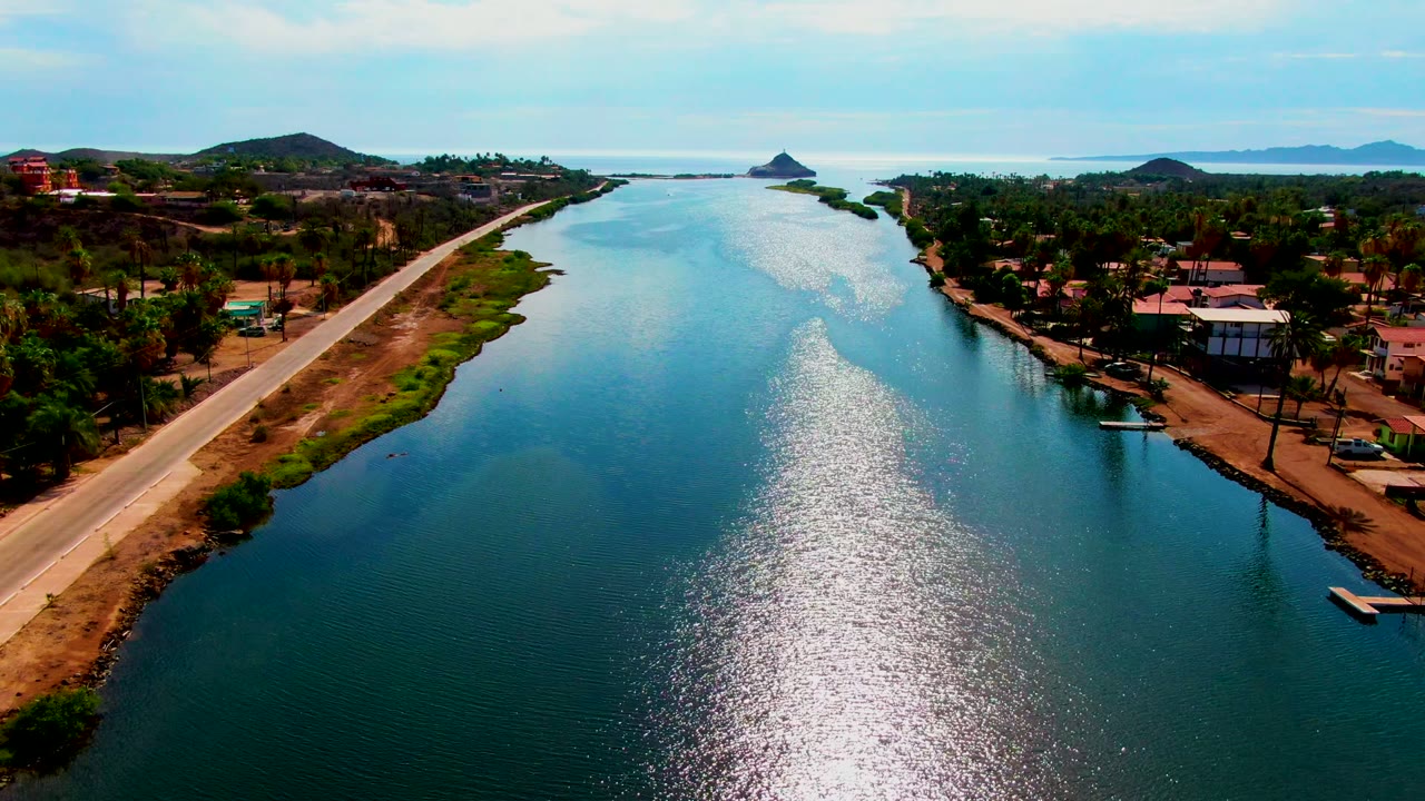 Drone view down a canal with a road on the left bank and buildings among trees on the right.