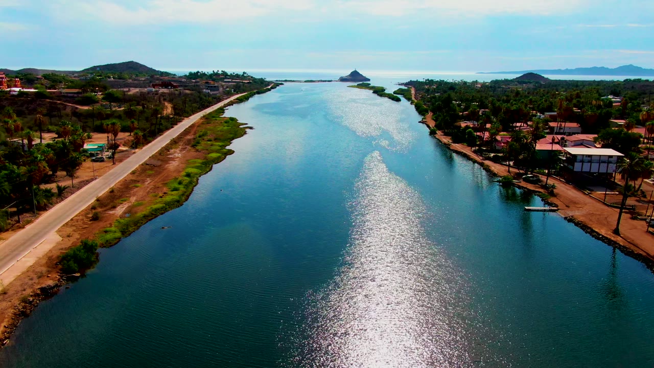 Mulege aerial frame showing shimmering blue-green canal water, a dry roadside bank on the left, clustered shoreline buildings and palms on the right, distant hills, and no visible people.