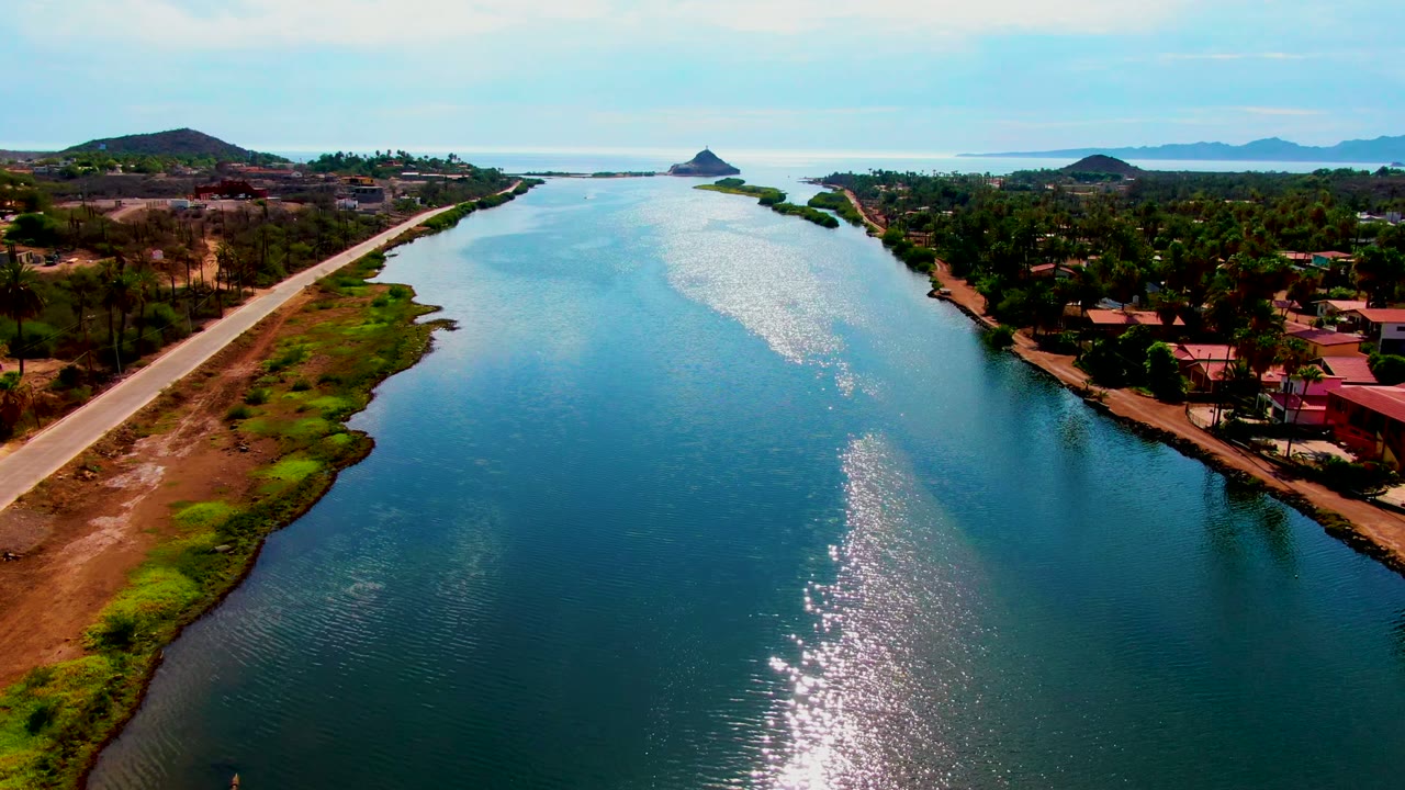 Opening aerial frame over a Mulege waterway, with sun reflection on blue water, a straight road along the left bank, trees and low buildings on the right, distant hills, and no visible people.