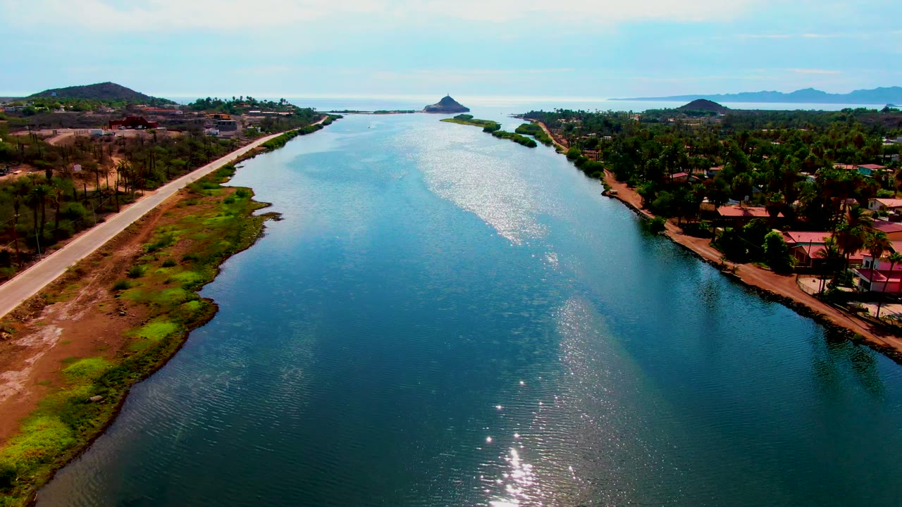 Aerial waterway scene with bright sun glint, shoreline vegetation, road, trees, and buildings