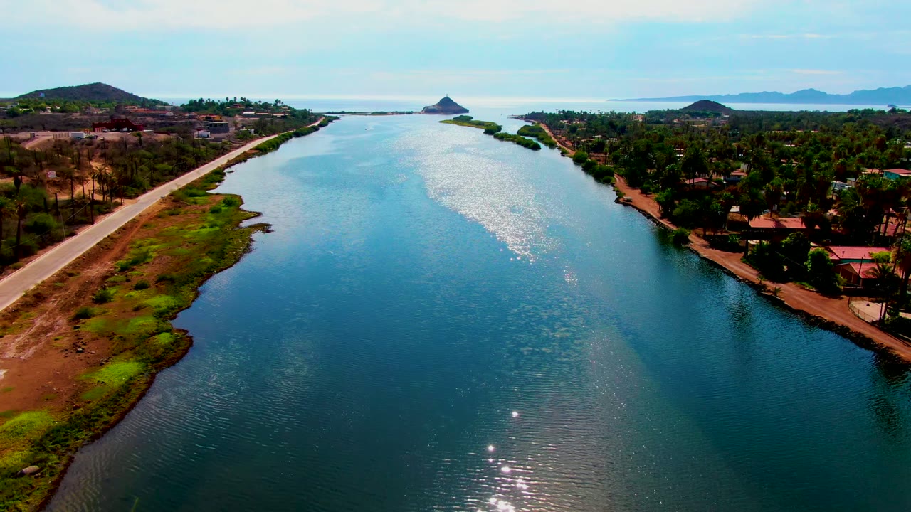At 0:02, the Mulege waterway remains centered with ripples catching light, a marshy left shoreline beside the road, dense trees and buildings to the right, hills on the horizon, and no visible people.