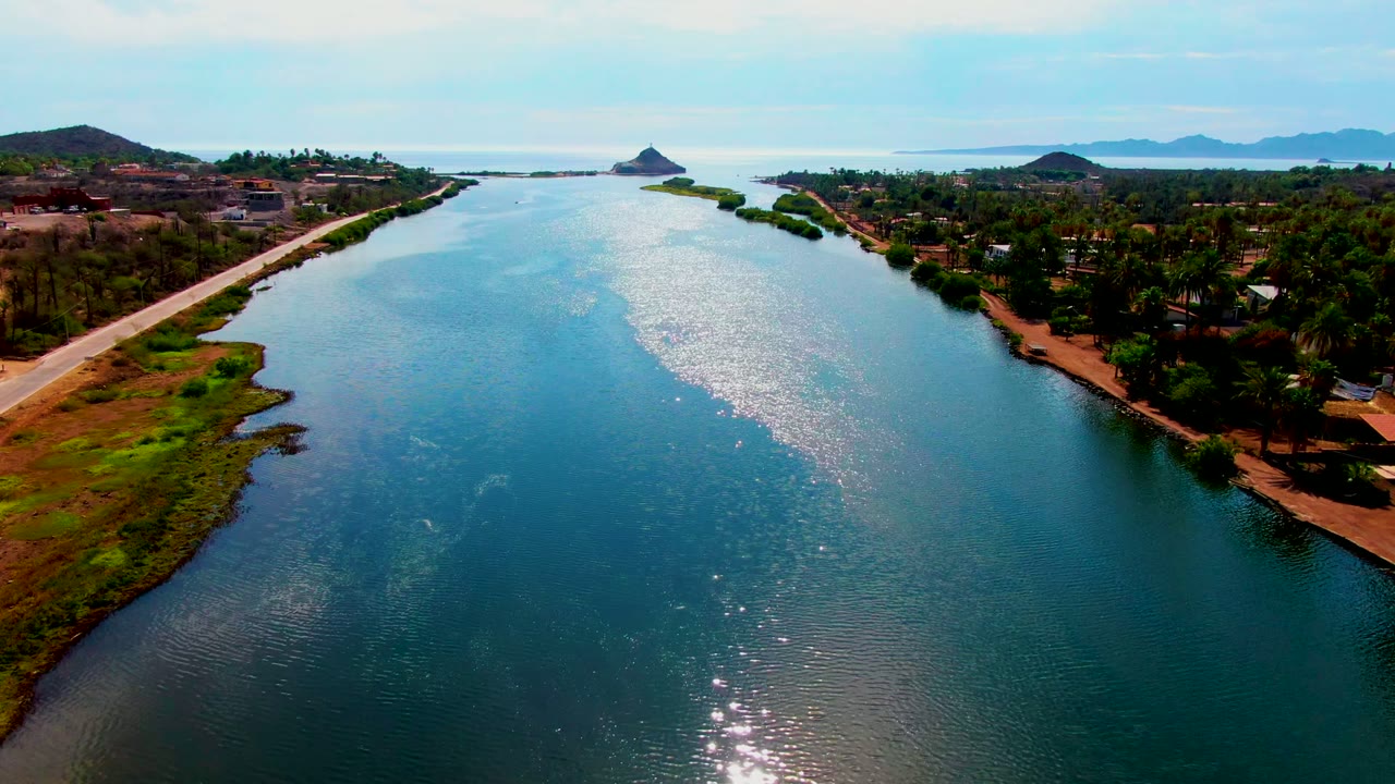 Opening aerial frame over the Mulege river in Baja California Sur, with blue-green water, road banks, palm trees, distant hills, and no visible people.