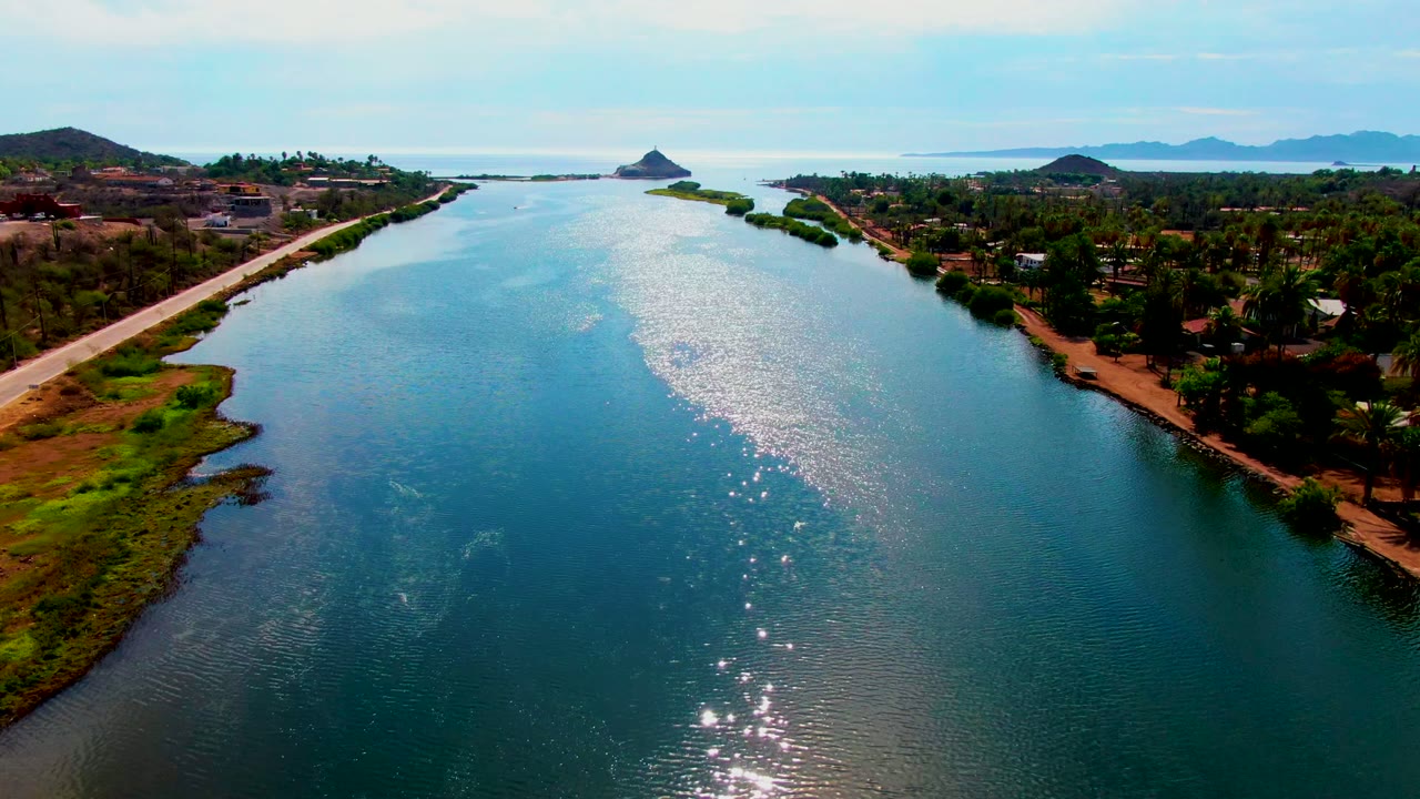 Aerial view along the Mulege river as the bright sun reflection stretches through the center water, with a straight roadside bank, palms, low buildings, and no visible people.