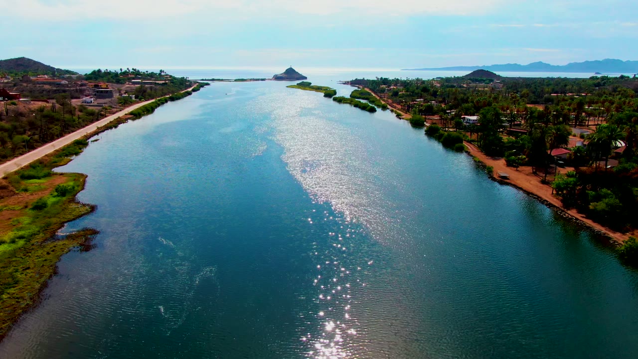 High aerial frame over the Mulege waterway, showing a marshy green edge in the lower left, a palm-lined settlement to the right, distant hills, and no visible people.