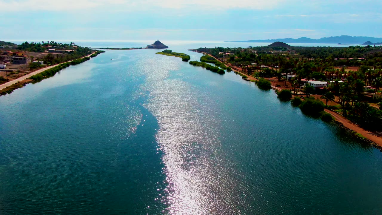 Opening aerial frame over a Mulege, Baja California Sur canal, with bright reflection on the blue-green water, palms and low buildings along the banks, and no visible people.