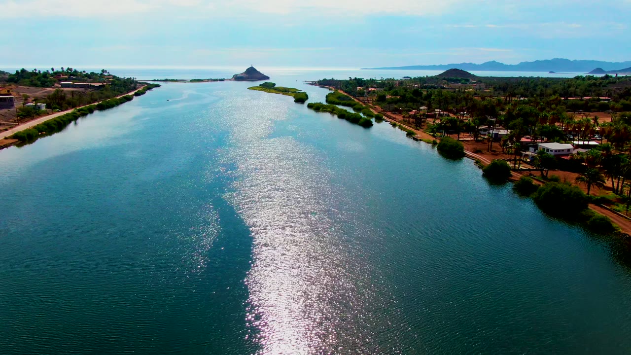 Mid-clip aerial view of the Mulege waterway, showing a wide band of glittering sunlight on rippled water, palms and small buildings near the shoreline, and no visible people.