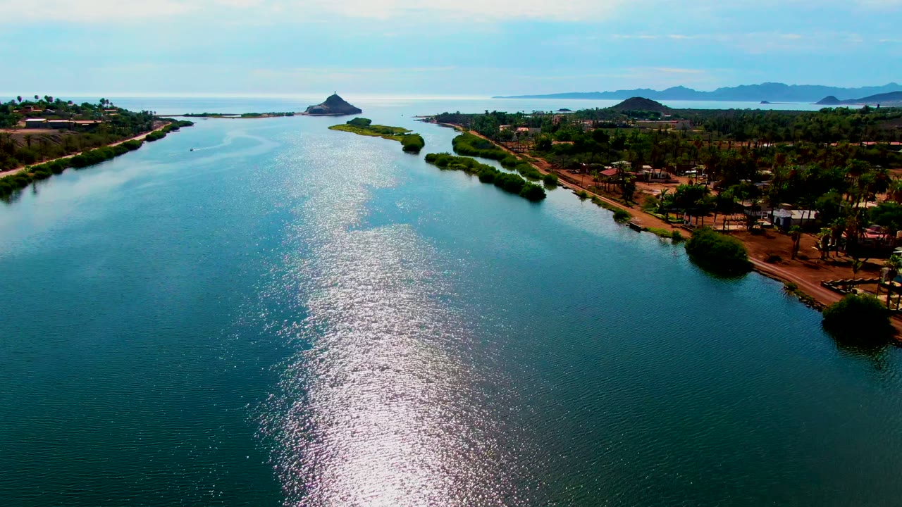 Opening aerial frame over a Mulege, Baja California Sur water channel, with a vertical path of sun glare on blue ripples, green shoreline trees, scattered buildings, distant hills, and no visible people.