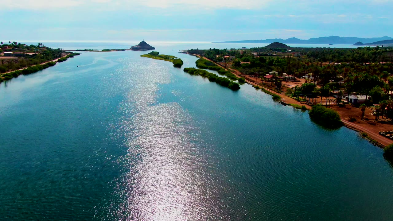 Drone view of sunlit channel water beside a tree-covered shoreline and dirt path.
