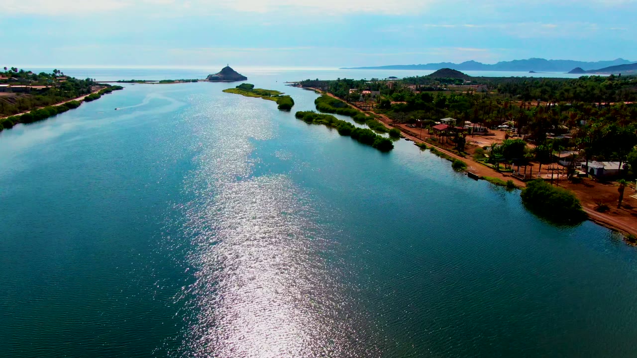 Drone frame over Mulege water with a bright reflective band across the channel, green banks on both sides, scattered low structures on the right shore, low hills beyond the water, and no visible people.
