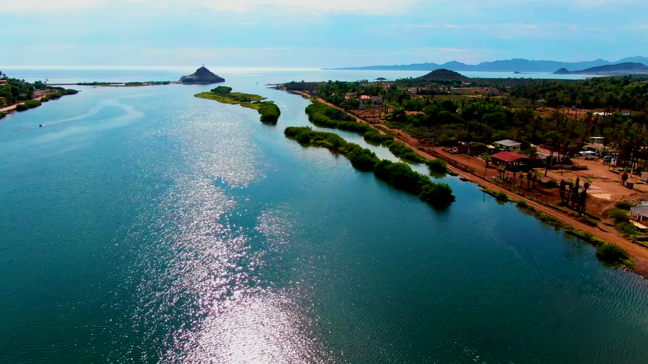 Aerial view in Mulege, Baja California Sur, Mexico, showing blue-green water with bright sun reflections, a vegetated shoreline, small buildings, distant hills, and no visible people.