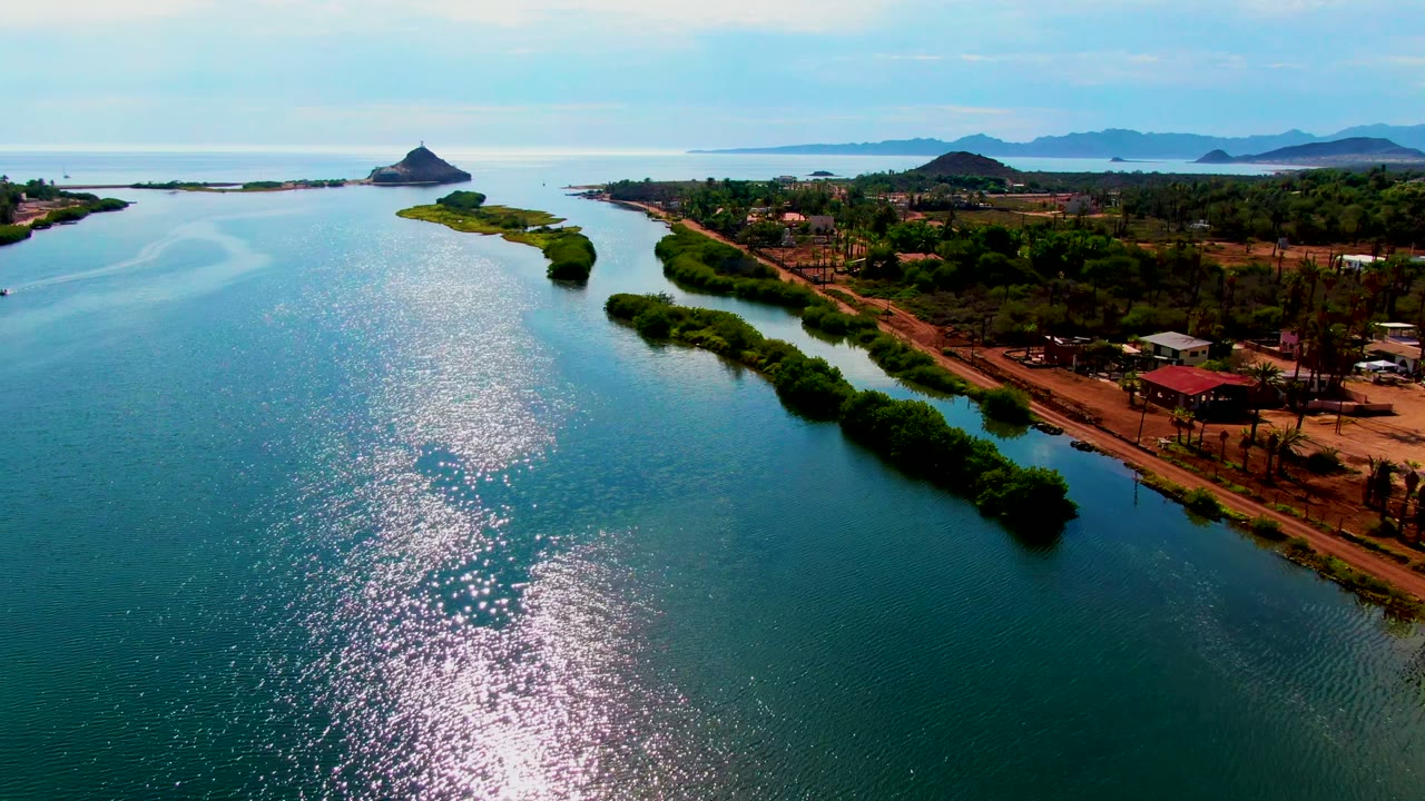 Wide aerial view in Mulege with shimmering blue water, narrow green shoreline vegetation extending into the channel, small buildings on land, distant hills, and no visible people.
