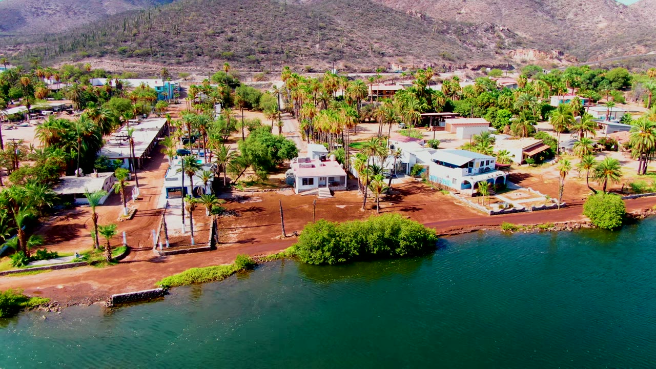 Aerial frame over Mulege, Baja California Sur, with green-blue water in the foreground, reddish shore, palm trees, small homes, dry hills in back, and no visible people.