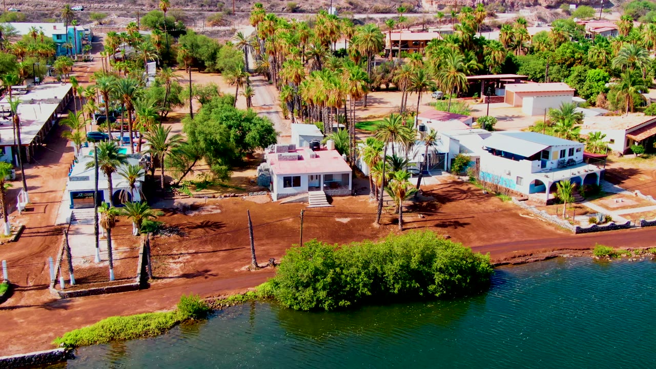 Opening aerial frame over Mulege, Baja California Sur, with green-blue water in front, a red-brown dirt shoreline, palms, low homes, and no visible people.