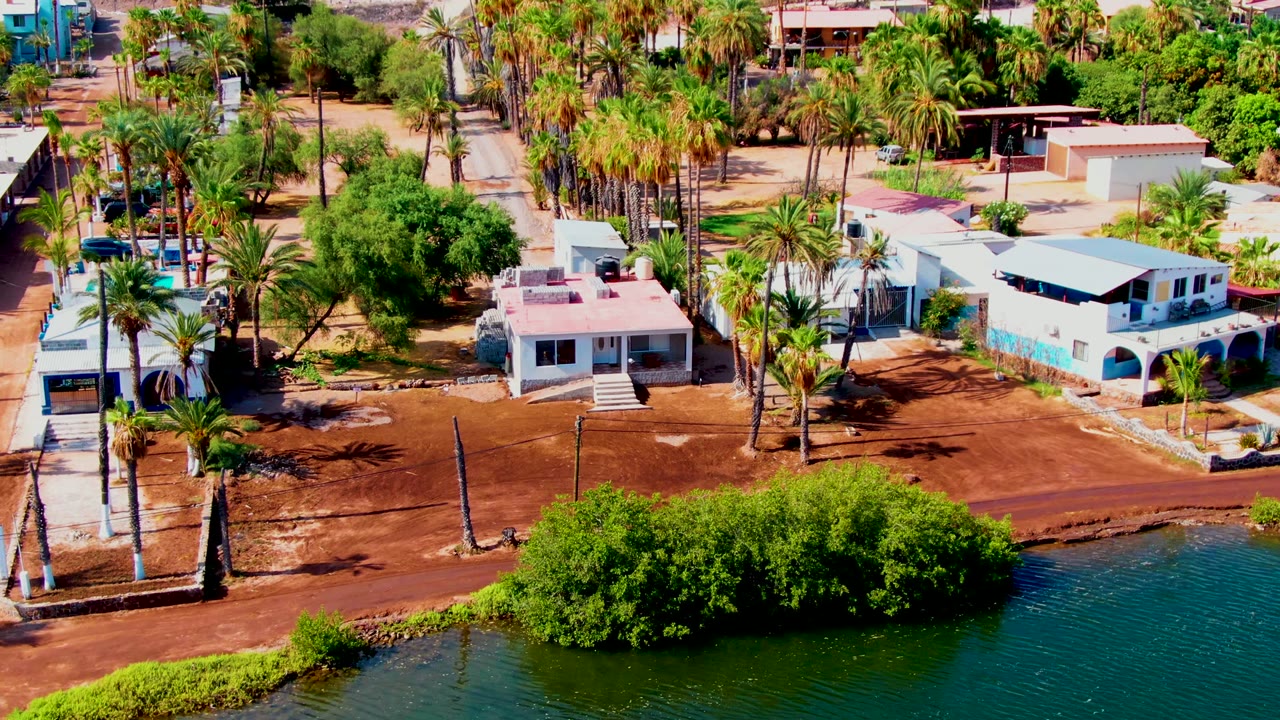 Mulege aerial frame with more of the right-side waterfront visible, including green-blue water, a red dirt road, palms, low houses, and no visible people.