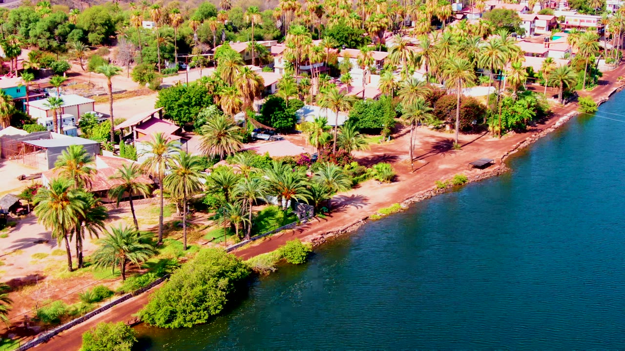 Opening aerial view in Mulege, Baja California Sur, with palm trees, small homes, reddish sandy shore, and calm blue water on the right, with no visible people.