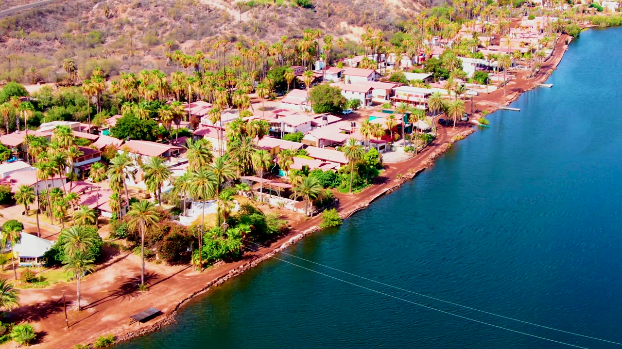 Aerial view in Mulege, Baja California Sur, showing palm-lined waterfront homes, reddish shoreline soil, dry hills, and blue water, with no visible people.