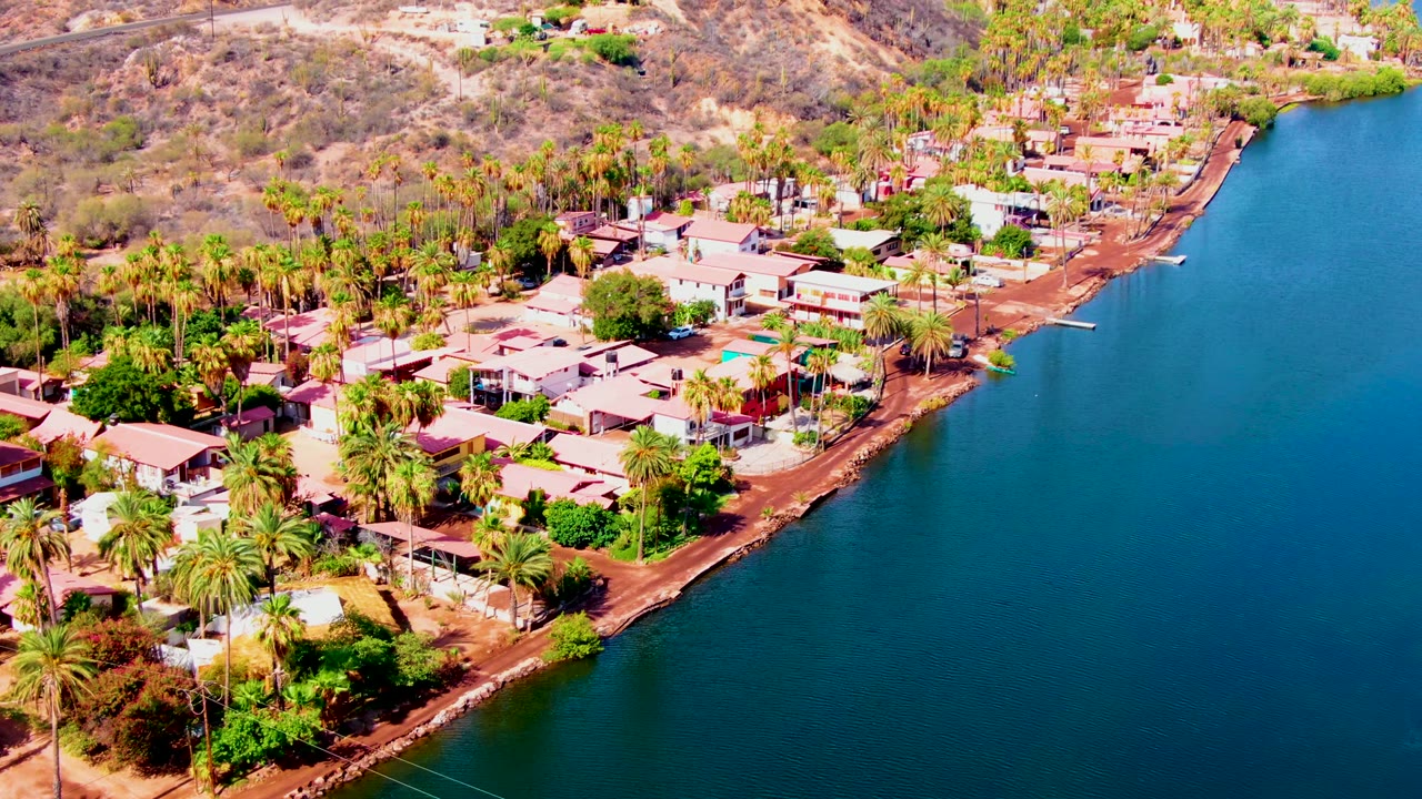 Aerial view in Mulege with a palm-filled residential strip below dry brown hills and a curved edge of dark blue water along the right side, with no visible people.