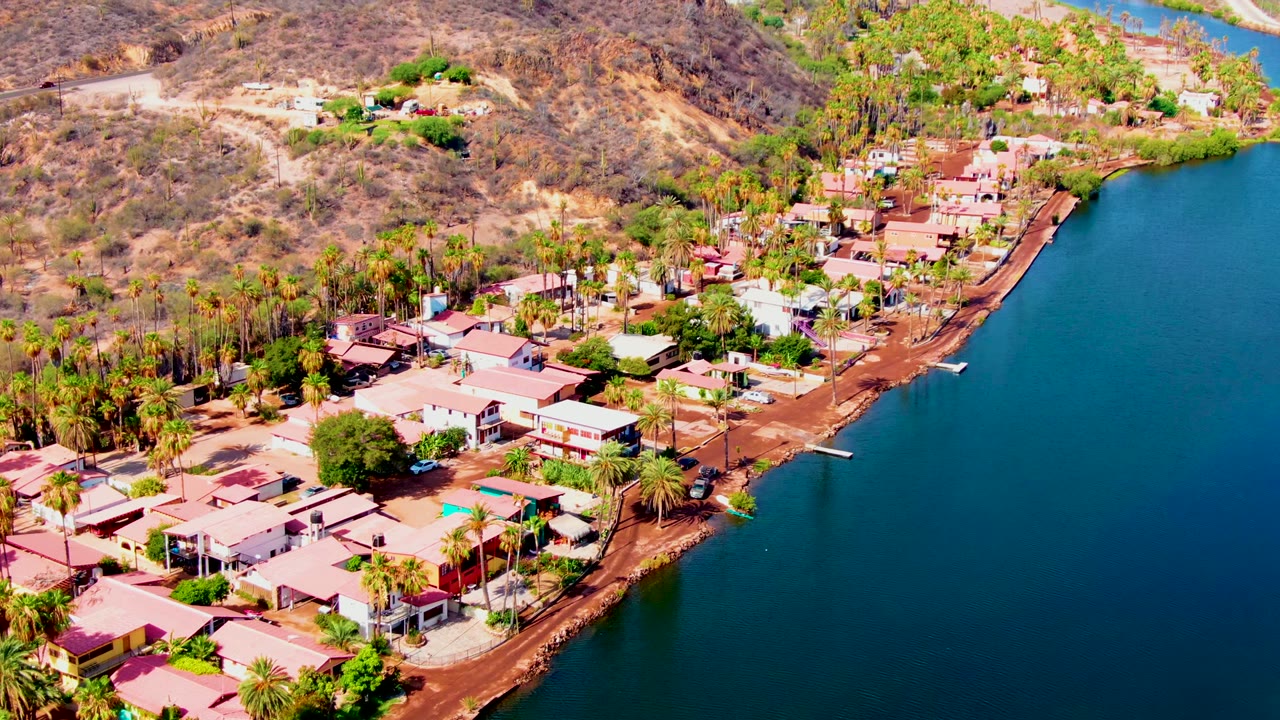 Aerial frame over Mulege, Baja California Sur, showing low homes, palm trees, reddish shore, dry hills, and blue water on the right, with no visible people.