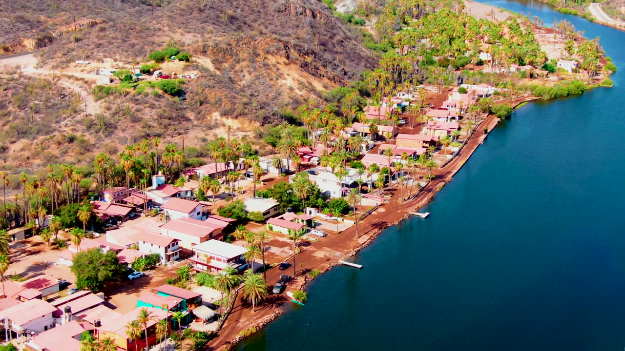 Mulege aerial frame with deep blue water dominating the right side, a reddish shore edge, palm trees, low houses, and arid hills beyond, with no visible people.