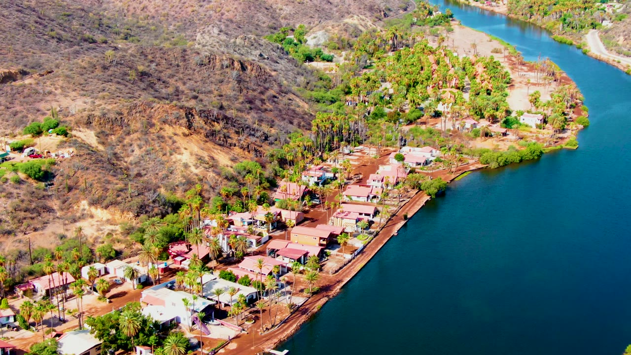 Aerial frame over Mulege, Baja California Sur showing lakeside homes, palms, a shoreline road, blue water on the right, dry brown hillside behind, and no visible people.