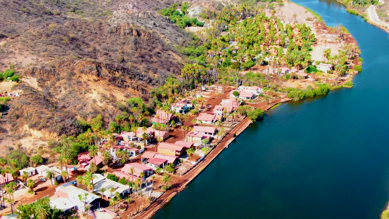 Aerial frame in Mulege, Baja California Sur with the blue waterway curving around a residential shore, palms and homes below, brown hillside at left, and no visible people.