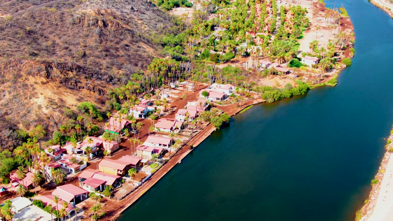 High aerial view over Mulege, Baja California Sur, with small homes along a blue-green lakeshore, palms and trees near red-brown soil, dry hills behind, and no visible people.