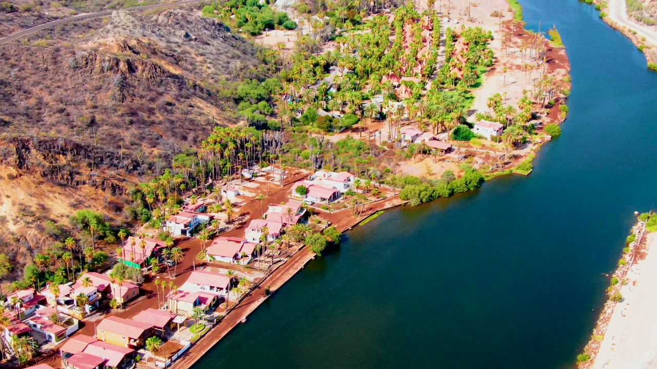 High-angle view in Mulege with residential roofs near the lower left, palms and green trees along the shore, dry hills in the upper left, calm water to the right, and no visible people.