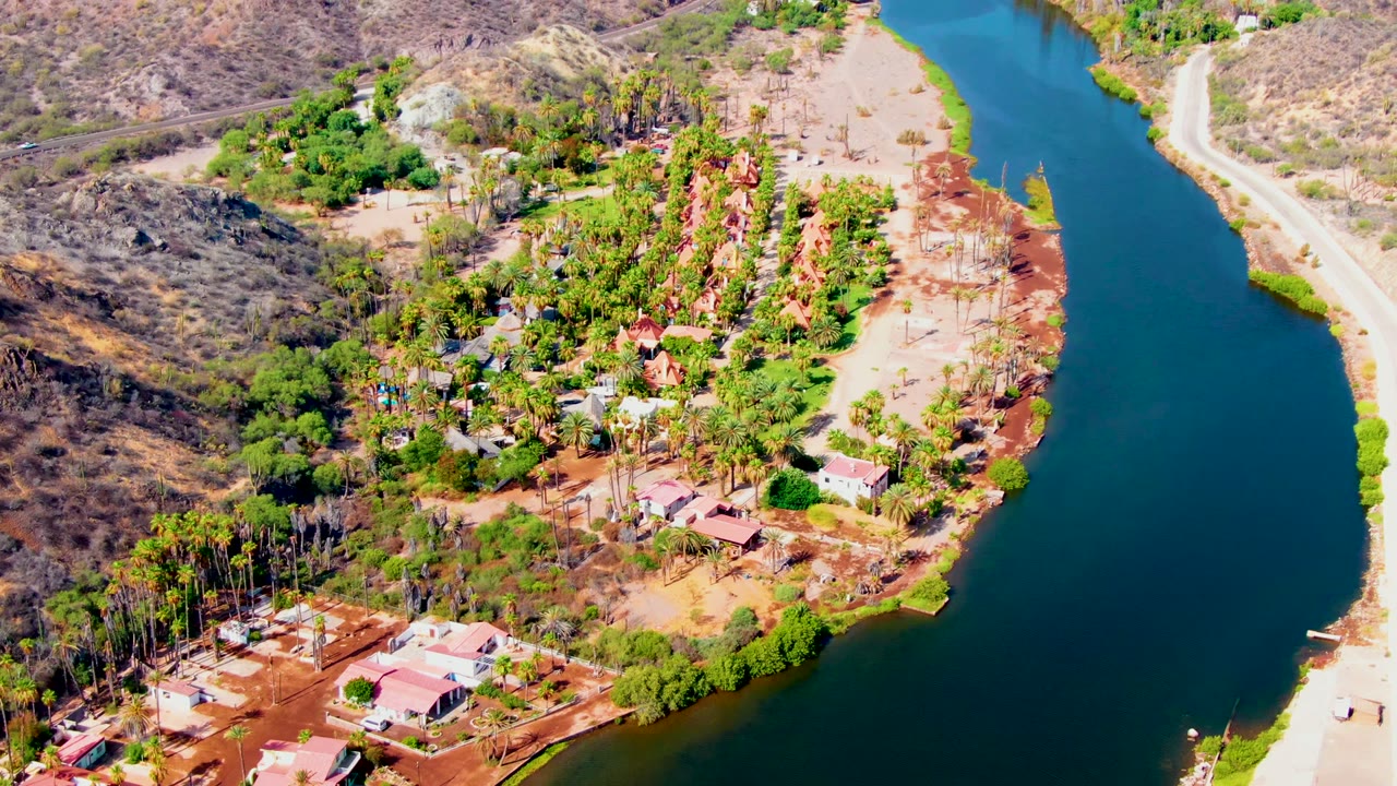 Aerial view in Mulege, Baja California Sur, showing a curving blue-green river, dry hills, scattered buildings, dense trees, and no visible people.