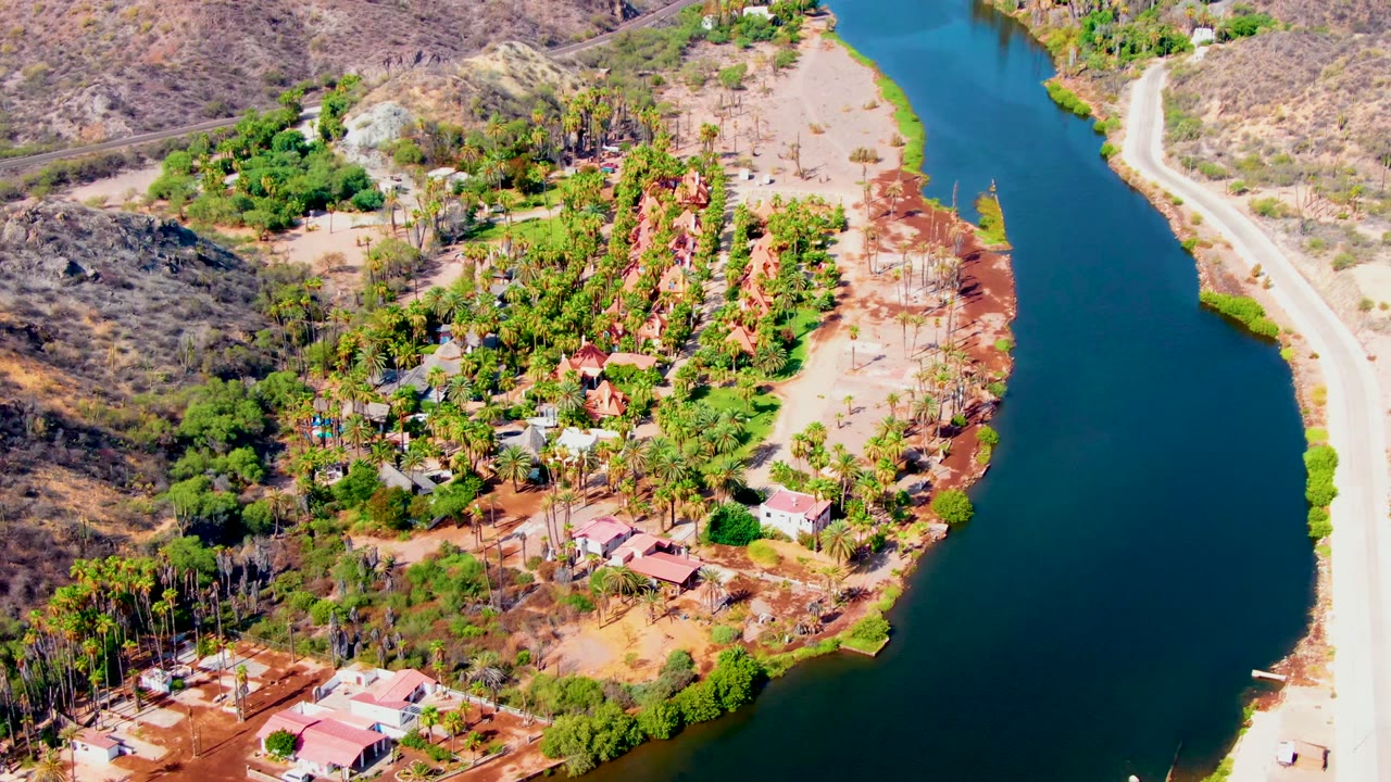 Aerial view of riverside homes, trees, a road, and hilly desert terrain.