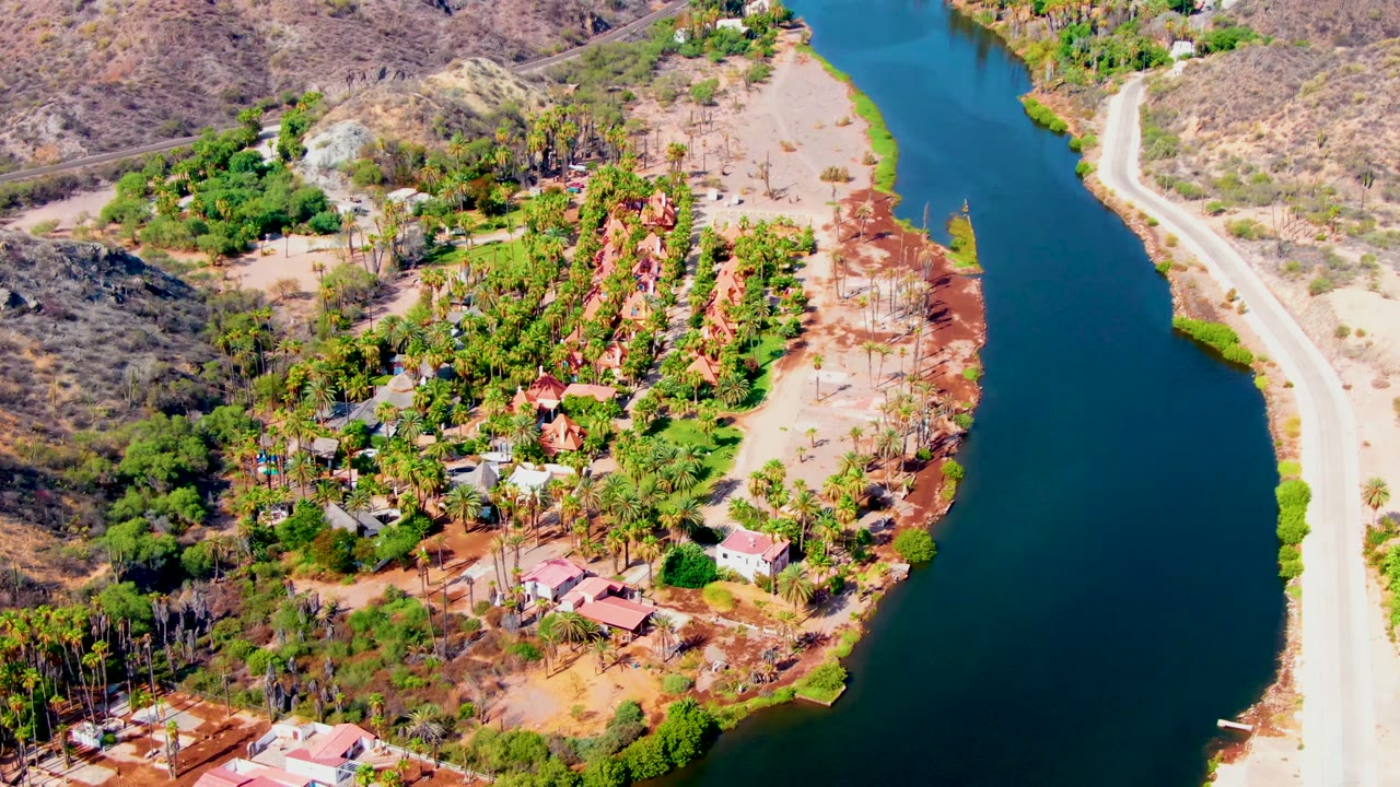 Aerial view in Mulege with the blue-green river running vertically through frame, a pale road on one bank, homes and trees on the other, dry hills, and no visible people.