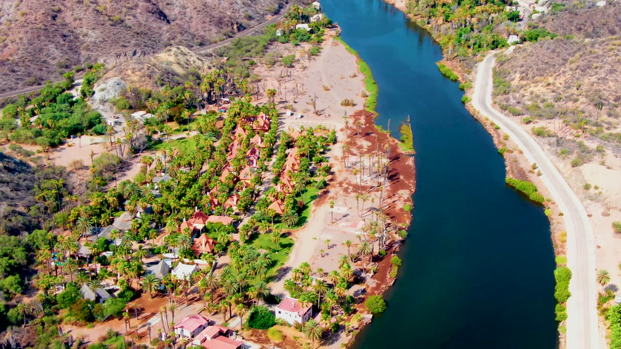 Aerial frame over Mulege, Baja California Sur showing a dark blue river canal, pale road, residential houses, palms, reddish soil, dry hills, and no visible people.