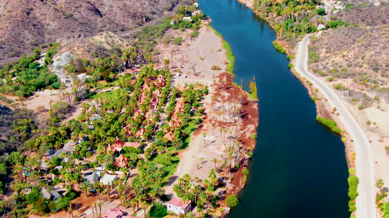 Drone view along a narrow waterway with a road on one bank and palms near houses.