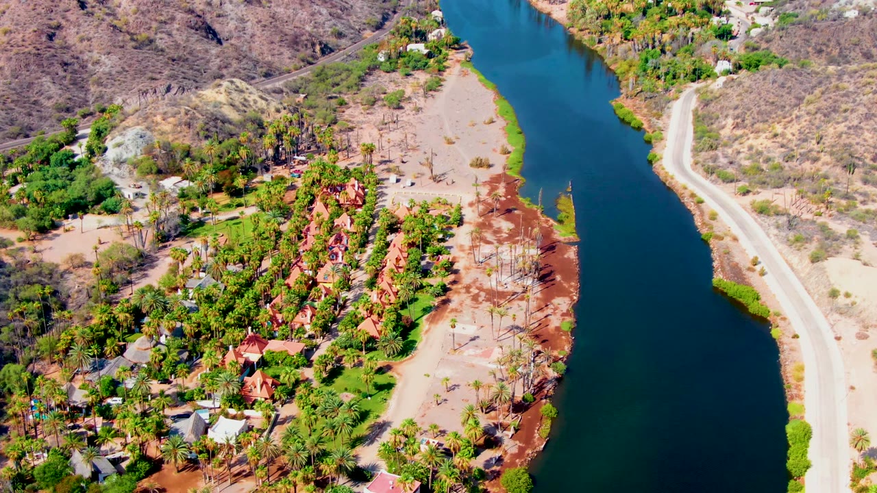 Mulege aerial frame showing the canal running vertically through the composition, a roadside bank on the right, clustered homes and palms on the left, reddish exposed soil, and no visible people.