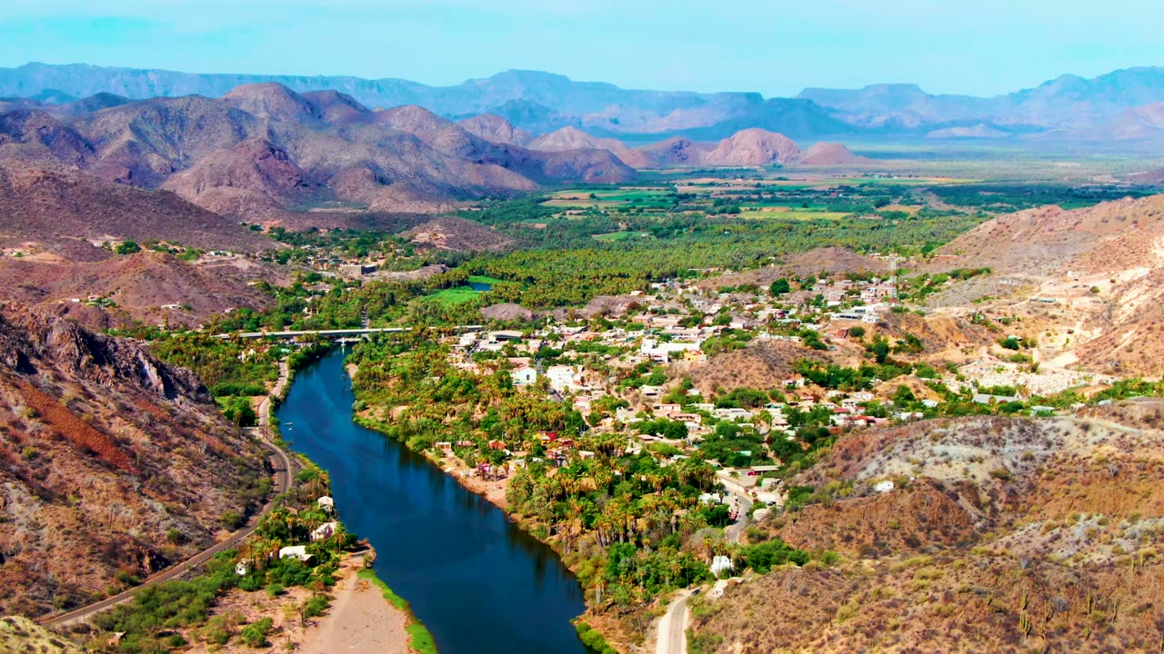 Aerial view over Mulege, Baja California Sur, with a blue river at lower left, green vegetation around town buildings, dry hills, distant mountains, and no visible people.