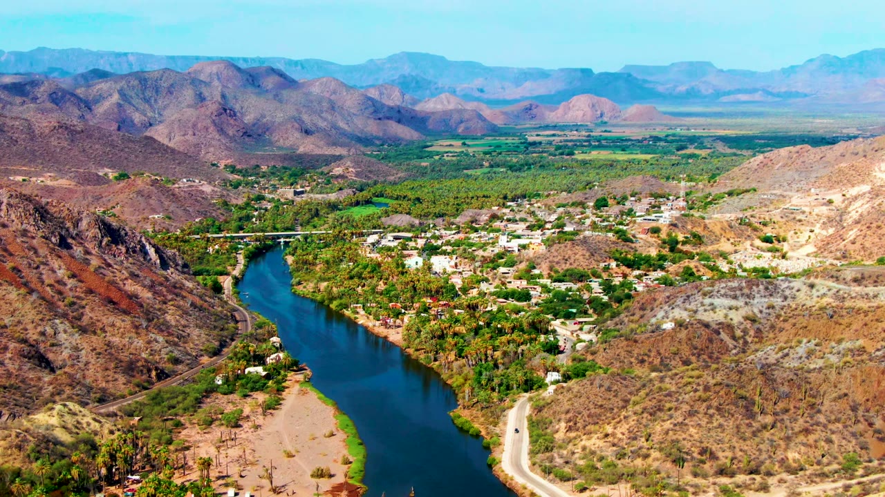 Opening aerial frame over Mulege, Baja California Sur, with a blue river bending through green palms, a pale road on the bank, clustered town buildings, dry hills, distant mountains, and no visible people.