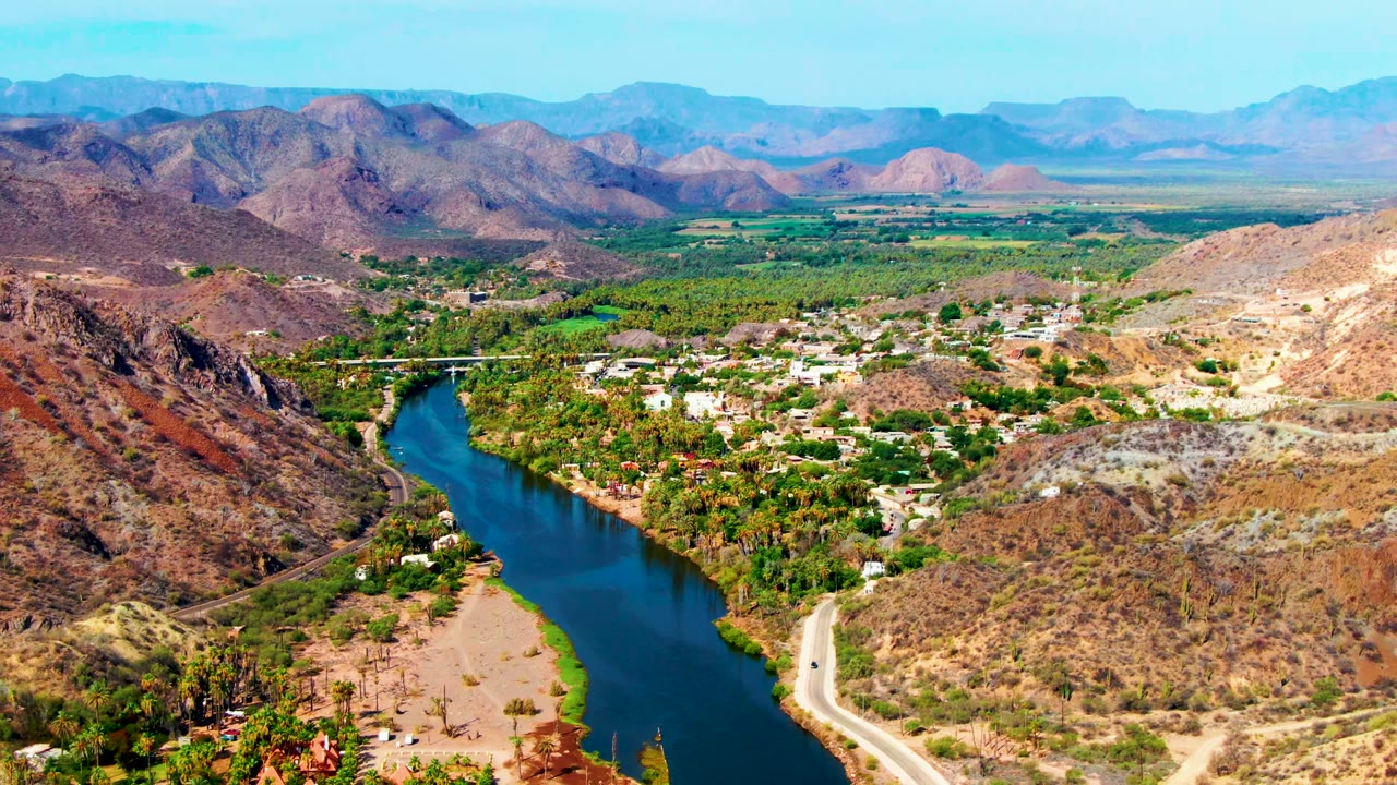 Mid-clip aerial frame in Mulege showing the blue river bordered by dense green vegetation, a light road on the right bank, town structures in the valley, brown hills, and no visible people.