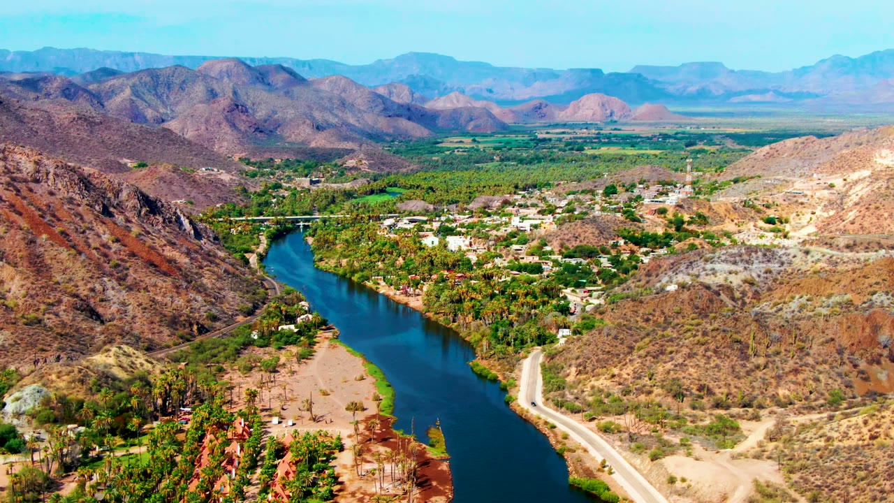 Wide aerial frame over Mulege, Baja California Sur, showing a blue river curving between arid hills, palms and trees along the bank, scattered buildings, distant mountains, and no visible people.