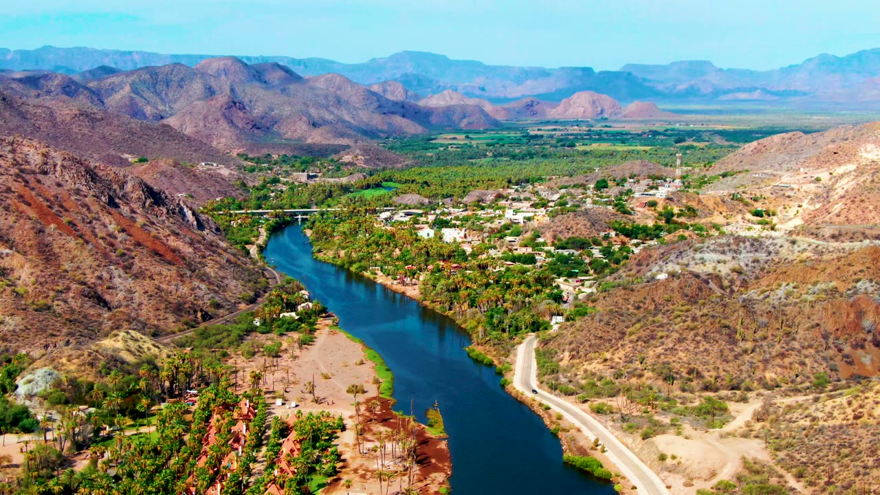 Aerial river valley with road, palms, dry hills, and distant mountains.