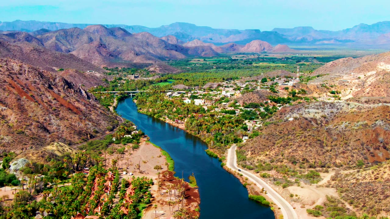 Drone-like aerial frame over Mulege, Baja California Sur, showing the river bend slightly right of center, a pale road beside it, clusters of trees and buildings in the valley floor, dry hills, and no visible people.