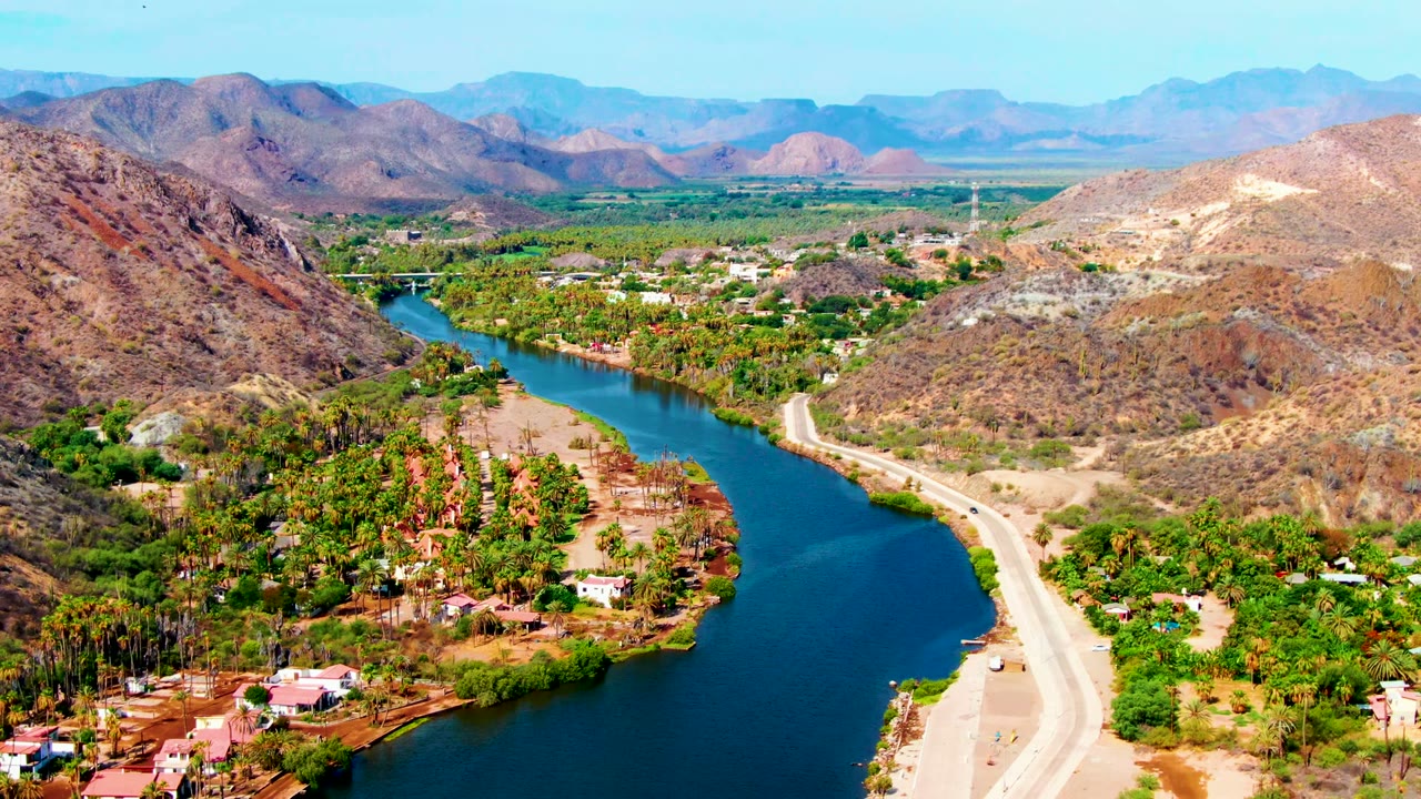 Aerial view over Mulege, Baja California Sur, with a blue river curving past green palms, a pale road, dry hills, distant mountains, and no visible people.