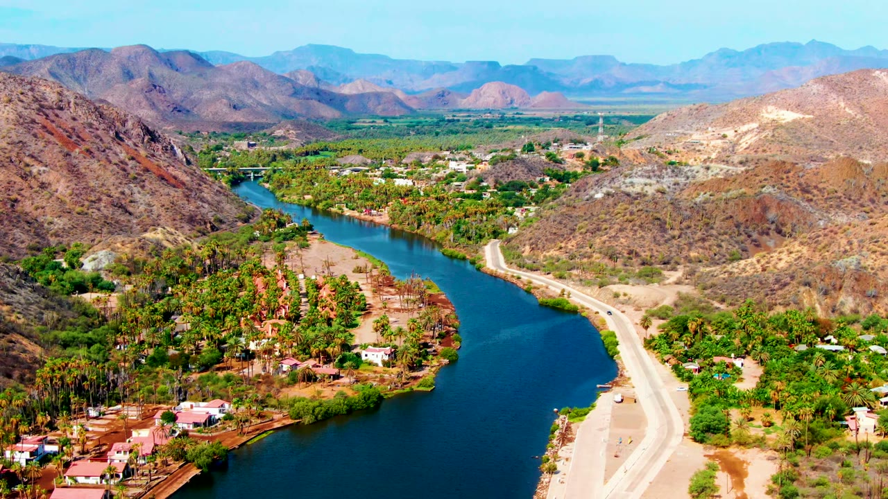 Wide aerial river valley with road, palms, houses, and brown hills under bright daylight.
