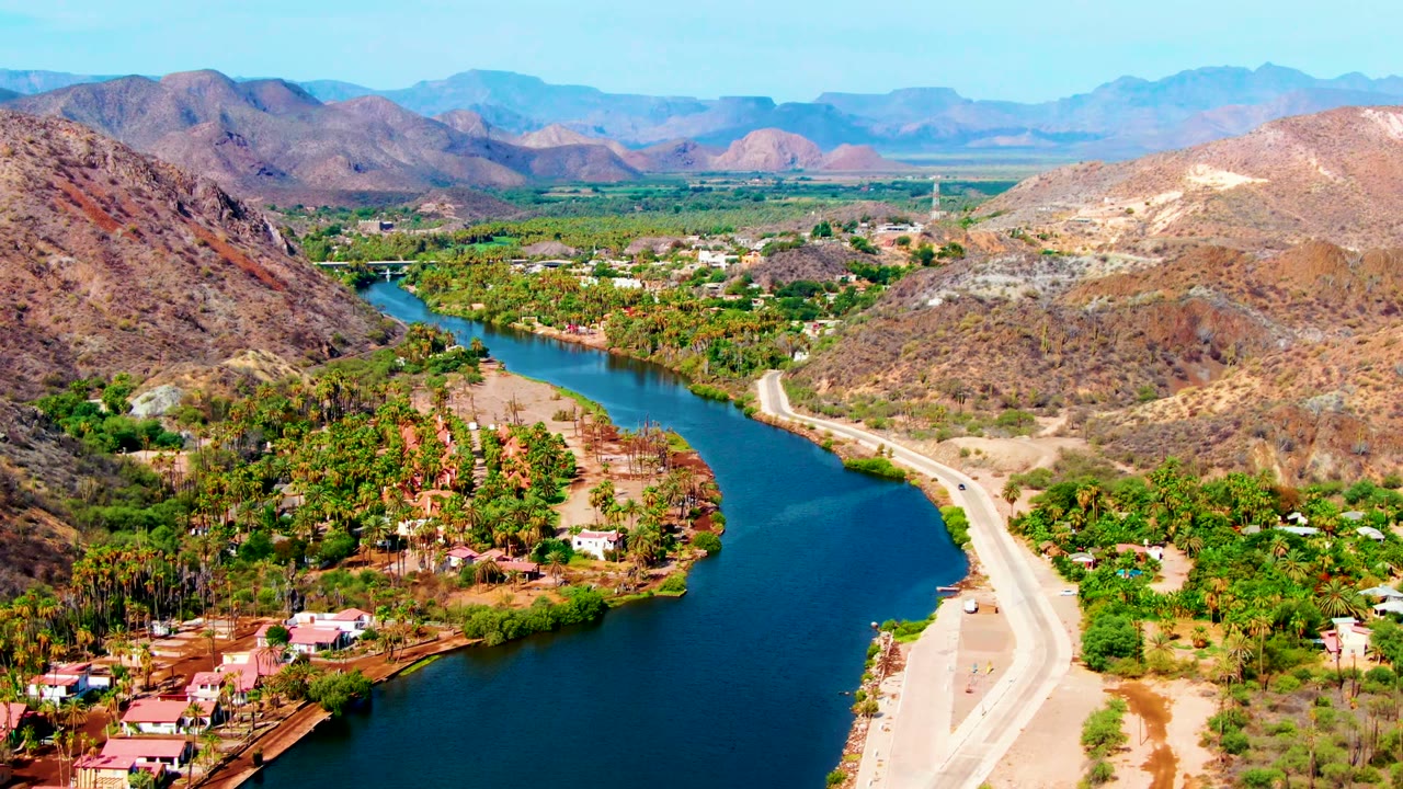 Aerial Mulege valley view with the dark blue river widening in the foreground, a light road tracing the bank, green trees around houses, and no visible people.