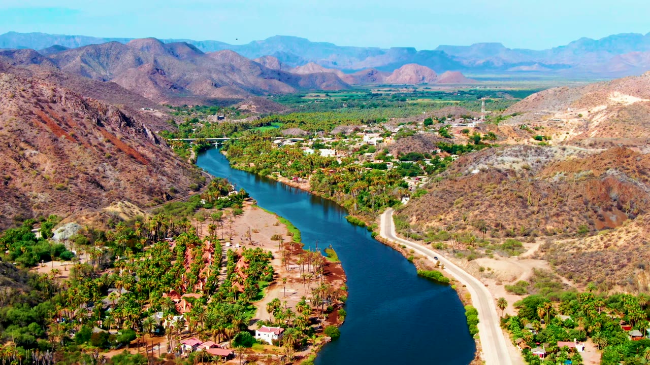 Aerial view over Mulege, Baja California Sur, with a blue river curving between dry hills, a pale road along the bank, scattered homes, dense greenery, distant mountains, and no visible people.