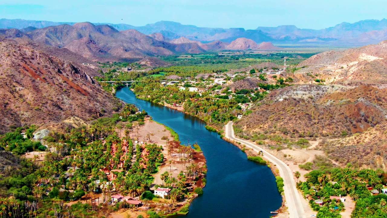 Wide aerial river valley with road, houses, greenery, and dry mountains.