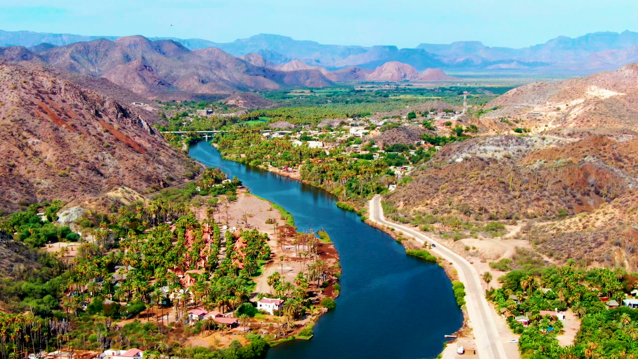 Aerial view in Mulege with the blue river cutting past a pale riverside road, green vegetation and low buildings on the valley floor, dry hillsides, distant mountains, and no visible people.