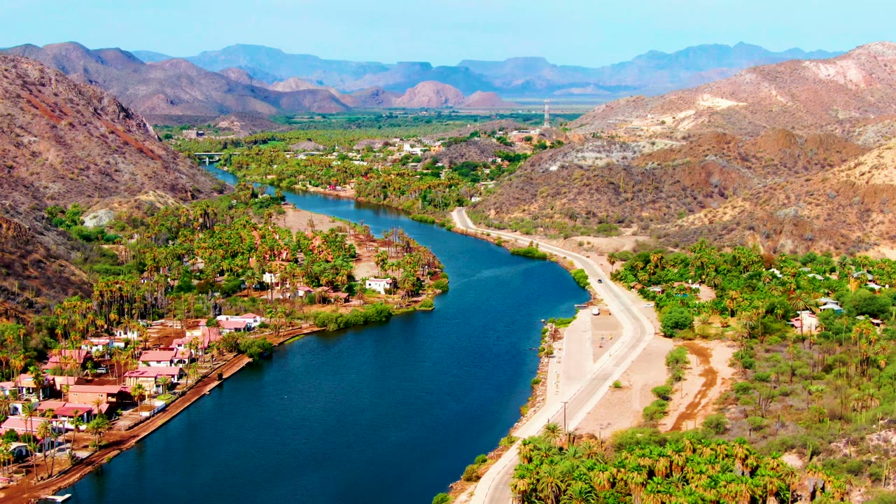 Opening aerial frame over Mulege, Baja California Sur, showing a blue river bend, palm-lined banks, scattered homes, a pale road, dry hills, distant mountains, and no visible people.